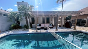 A swimming pool with a wooden deck and lounge chairs, surrounded by palm trees and a covered patio area. - Padre Island house with private pool (Corpus Christi)