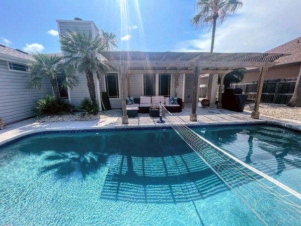 A swimming pool with a wooden deck and lounge chairs, surrounded by palm trees and a covered patio area.