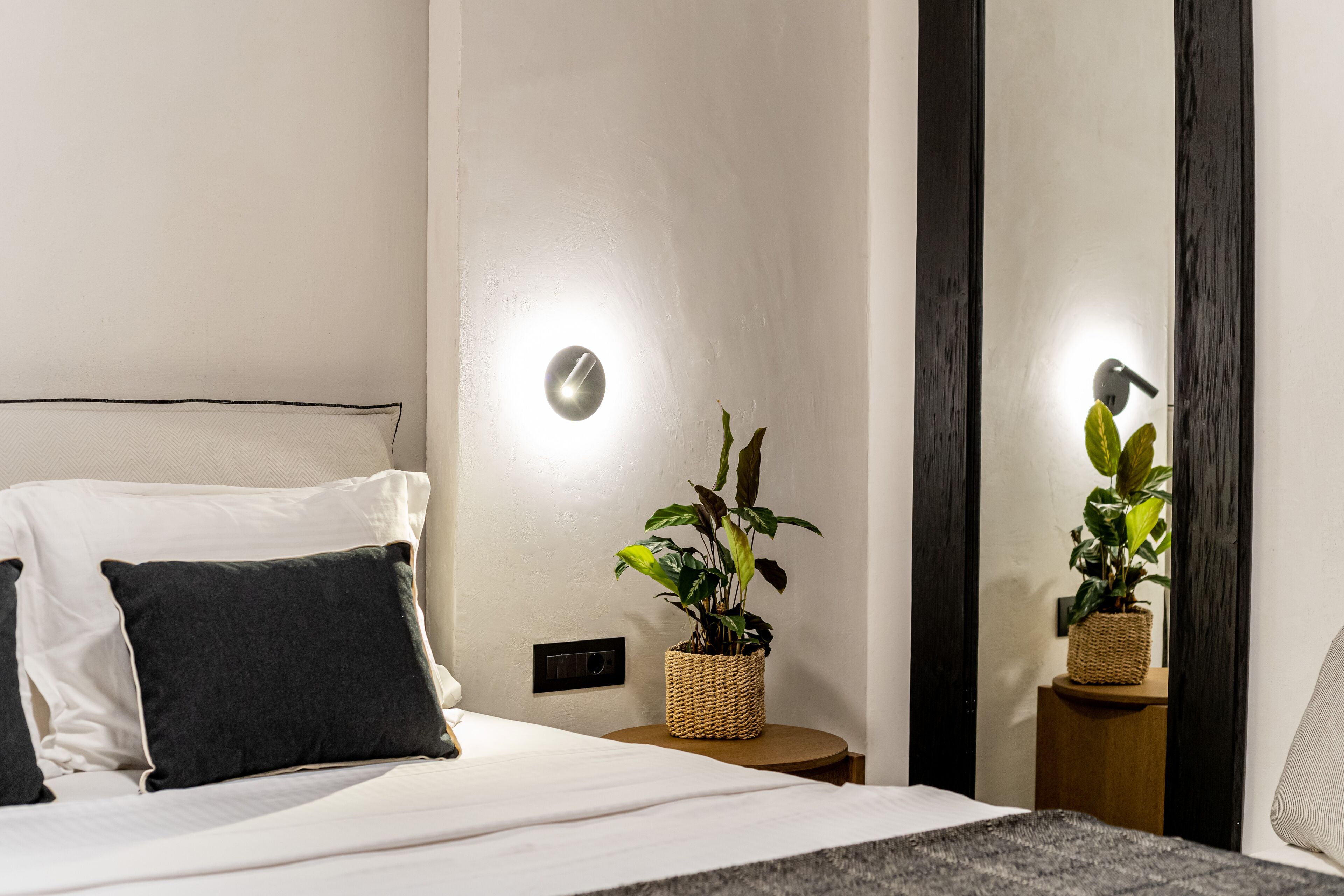 A neatly made bed with white and dark pillows, a bedside plant in a woven basket, and a wall-mounted light.