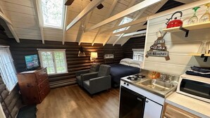 A cozy cabin-style room with a loft bed, a small kitchenette, a fireplace, and a window overlooking the forest.