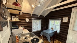 A compact kitchen with a sink, stove, and wooden walls. There are two windows with blinds, a table with chairs, and a clock on the wall.