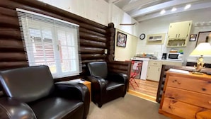 Una habitación acogedora con una pared de troncos, dos sillones de cuero, un aparador de madera y una kitchenette al fondo.