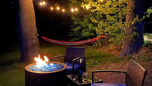 Un pozo de fuego con llamas, rodeado de sillas y una mesa, con árboles y luces colgantes de fondo.