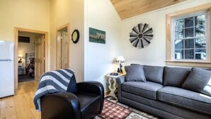Una sala de estar acogedora con techo de madera, un sofá gris, un sillón negro y una mesita con una lámpara.