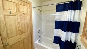Un baño con puerta de madera, ducha con cortina azul y blanca, y bañera blanca.