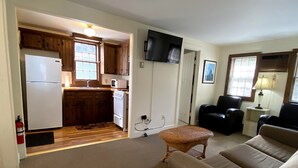 A compact living space with a kitchenette, a flat-screen TV mounted on the wall, a sofa, and a chair.