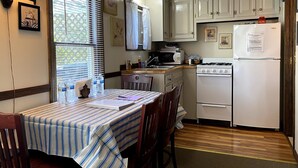 A compact kitchen with white cabinets, a stove, a microwave, and a refrigerator. There is a dining table with chairs and a window with blinds.