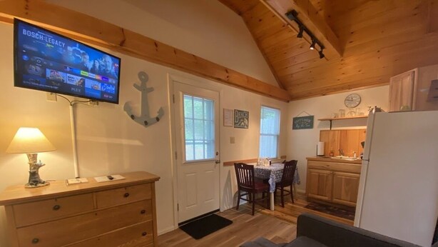 A cozy cabin interior with a wooden ceiling, a television mounted on the wall, a dining area with a table and chairs, and a kitchen with wooden cabinets.