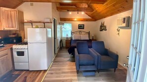 A compact kitchen with a stove, refrigerator, and wooden cabinets. A blue sofa is positioned in the living area, and a bed is visible in the background.
