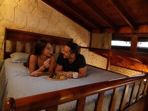 Um casal sentado na cama, com uma parede de pedra ao fundo, um corrimão de madeira na frente e vista para o exterior.