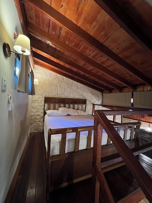 Techo de madera, pared de piedra, cama con ropa blanca, mesita de noche y una lámpara.