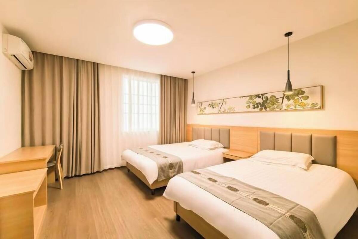 A hotel room with two beds, a desk, a chair, a ceiling light, and a wall-mounted artwork.