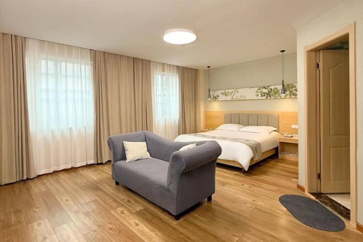 A hotel room with a bed, a sofa, a bedside table, a lamp, and a large window with curtains.
