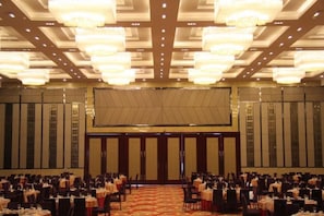 A banquet hall with rows of tables and chairs, a stage at the front, and chandeliers.