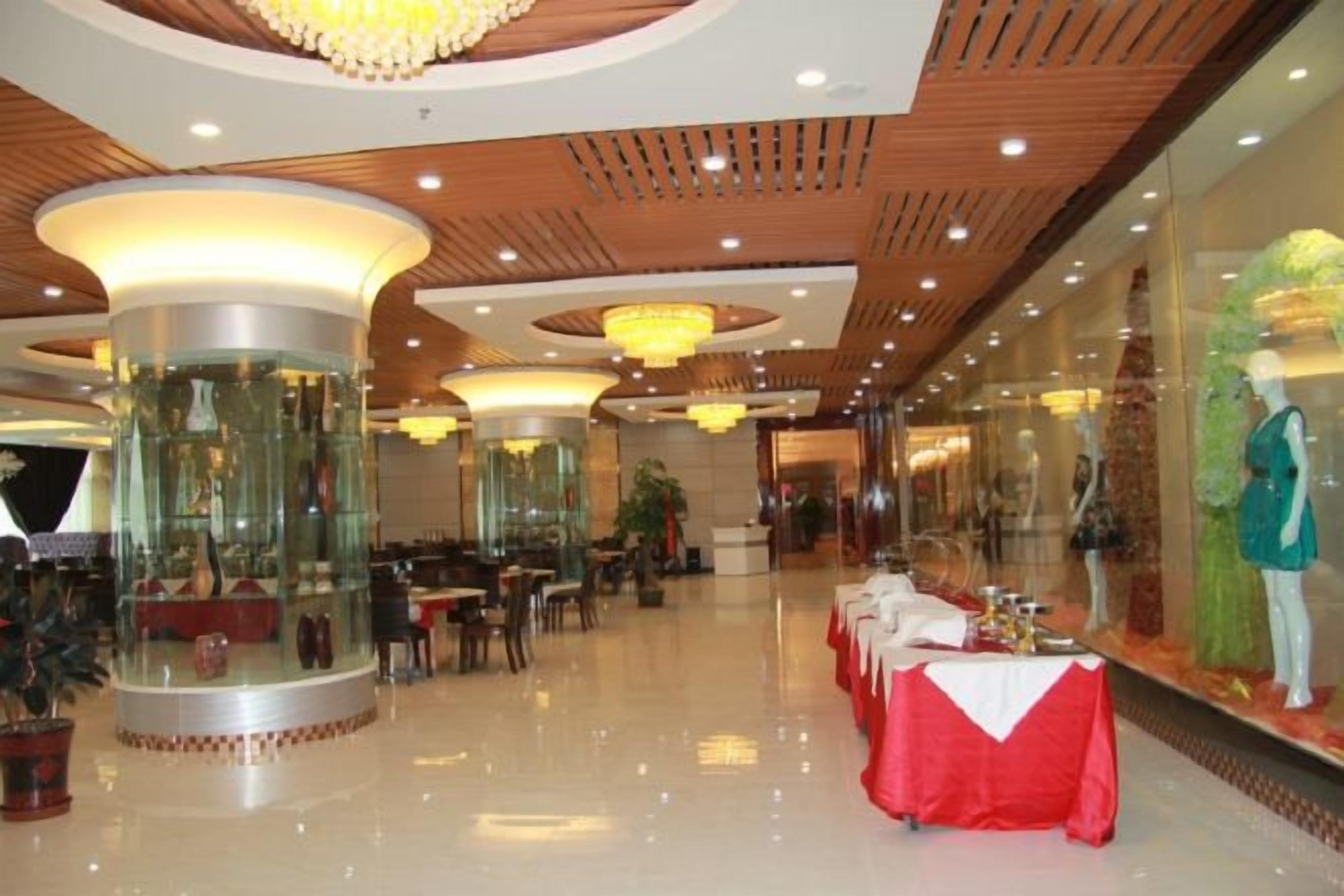 A spacious hotel lobby with high ceilings, large chandeliers, and a central column. Tables are set up for dining, and there are mannequins on display.