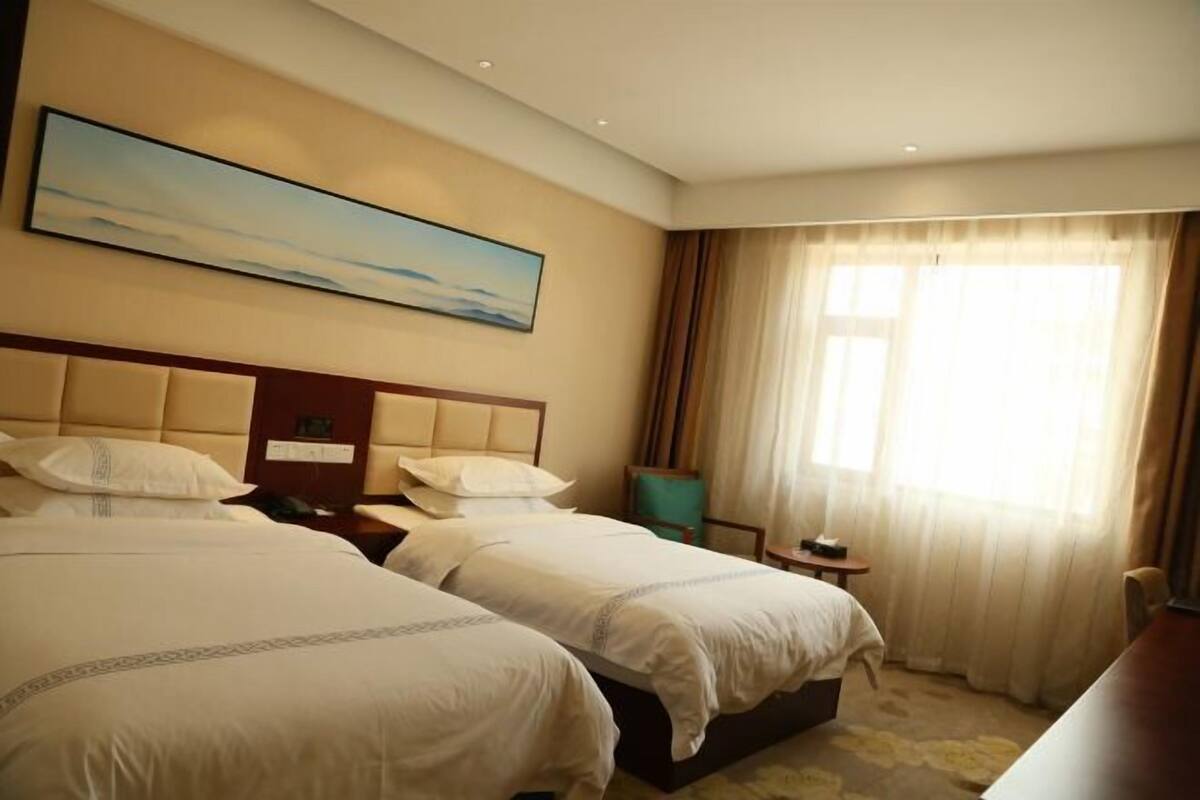 Une chambre d’hôtel avec deux lits, une tête de lit, un tableau, une fenêtre avec des rideaux, une petite table et une chaise.