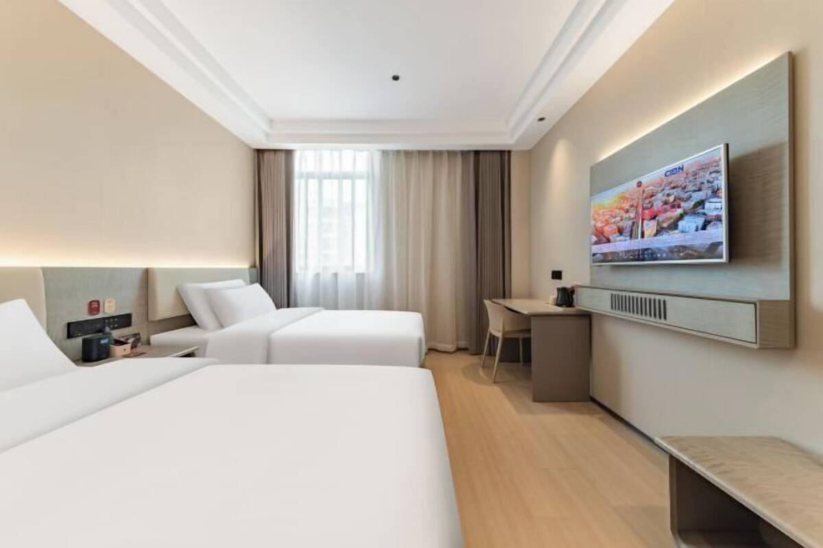 Quarto de hotel com duas camas, uma escrivaninha, uma cadeira, televisão e janela com cortinas.