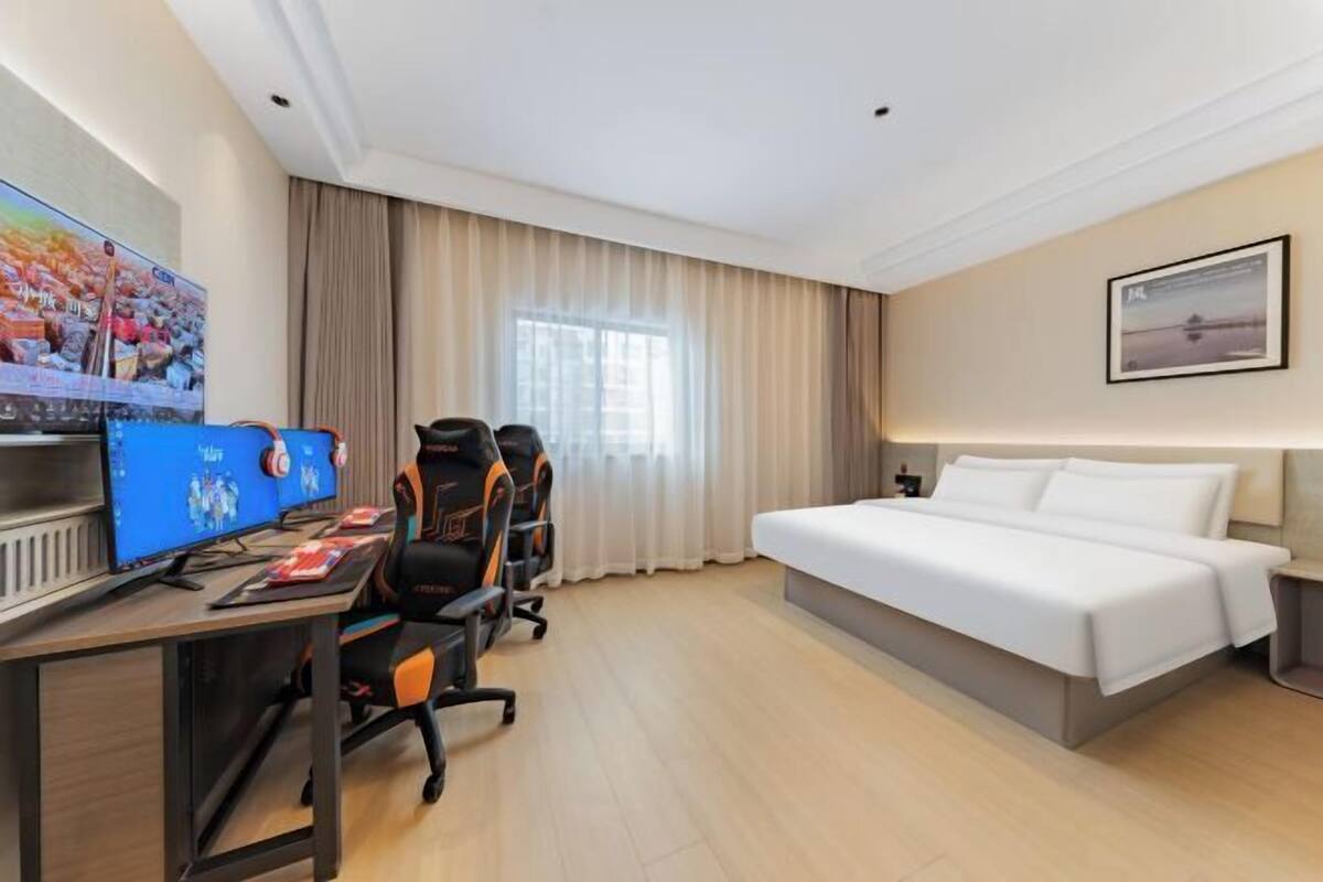 Quarto de hotel com cama, escrivaninha com dois monitores, cadeiras gamer, televisão e janela com cortinas.