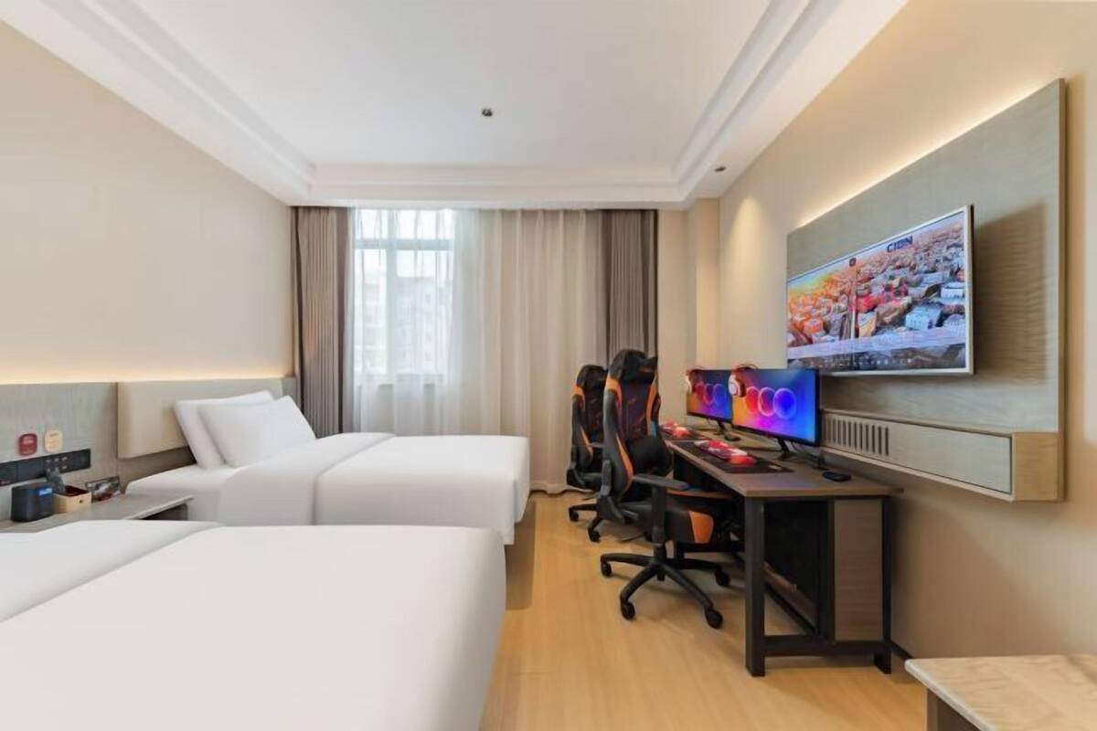 Quarto de hotel com duas camas, uma escrivaninha com computador, televisão e janela com cortinas.