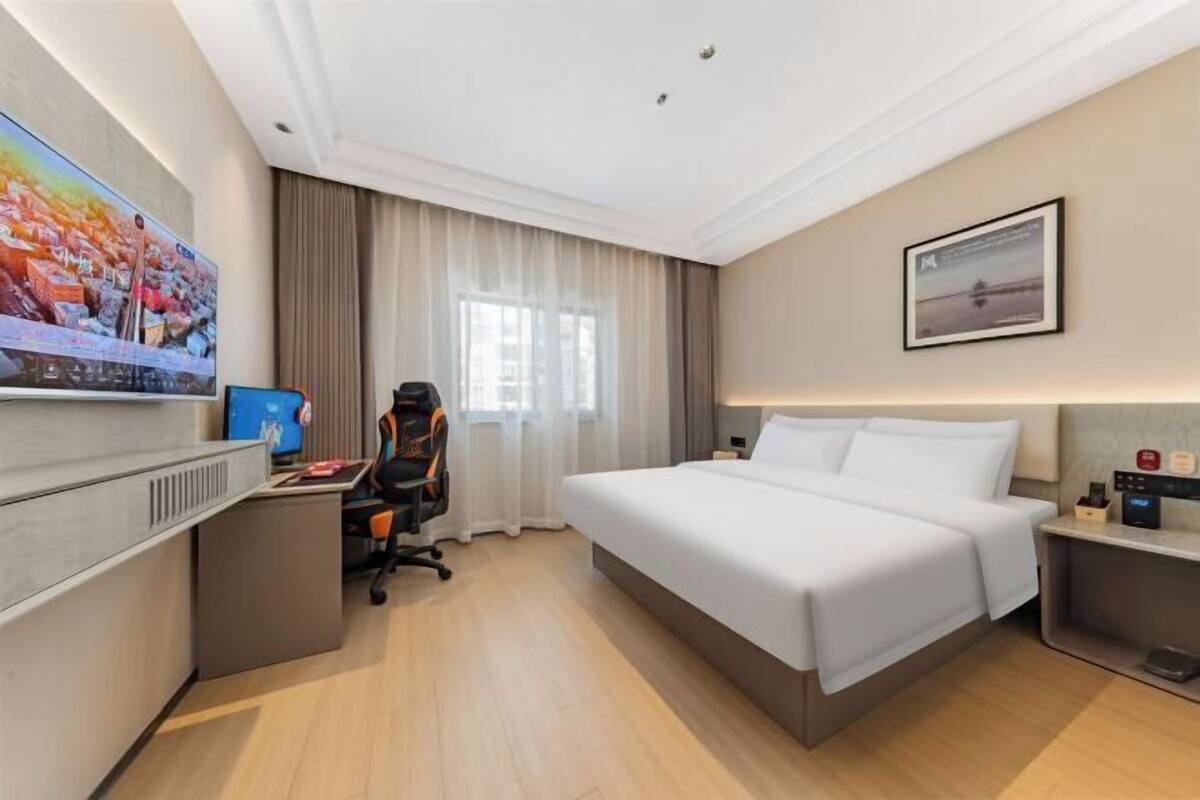 Quarto de hotel com cama, escrivaninha com computador, cadeira, televisão e um quadro na parede.