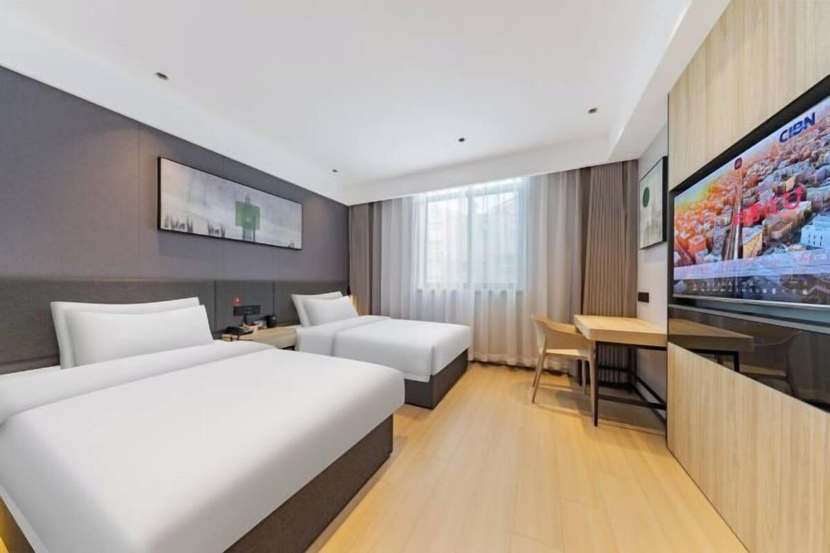 Quarto de hotel com duas camas, uma escrivaninha, televisão e uma janela grande com cortinas.