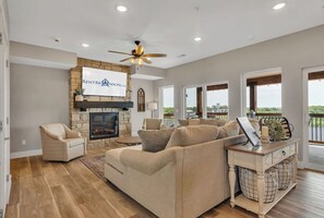 A spacious living room with a stone fireplace, a ceiling fan, and a sign for 'RENTBR' on the wall. - Lakehouse Lodge w/ Custom Decor & Outdoor Living Area, Hot Tub, Lake Views, Billiards & Free Tickets (Branson)