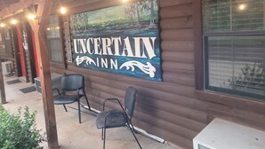 A wooden exterior wall with a 'Welcome' sign and a large 'Uncertain Inn' sign. There are two black chairs outside. - Fun In The Sun #11 (Karnack)