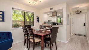 A dining area with a wooden table and chairs, a blue armchair, a window with a view of greenery, and a kitchen in the background. - 1/2 Mile to Beach, 2 bedroom townhome close to Mayo Clinic (Jacksonville Beach)