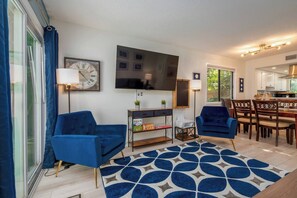 A living room with two blue armchairs, a coffee table, a TV, a clock, and a dining area with wooden chairs and a table.