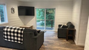 A living room with a dark grey sofa, a smaller armchair, and a coffee table. There is a flat-screen TV mounted on the wall, and a sliding glass door leading to an outdoor area. - Hidden Hideway, fully furnished home perfect for family get aways! (Boyne City)