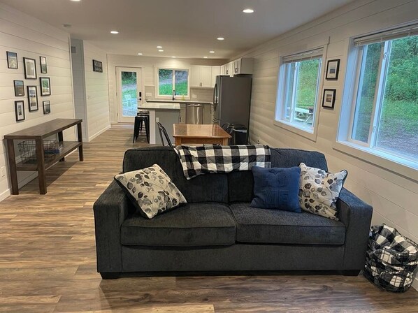 A modern living room with a dark grey sectional sofa, a wooden coffee table, and a black and white checkered throw. A kitchen with white cabinets and a stainless steel refrigerator is visible in the background. - Hidden Hideway, fully furnished home perfect for family get aways! (Boyne City)