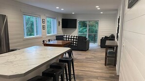 A modern kitchen with a large island, bar stools, a flat-screen TV, and a view of the outdoors. - Hidden Hideway, fully furnished home perfect for family get aways! (Boyne City)