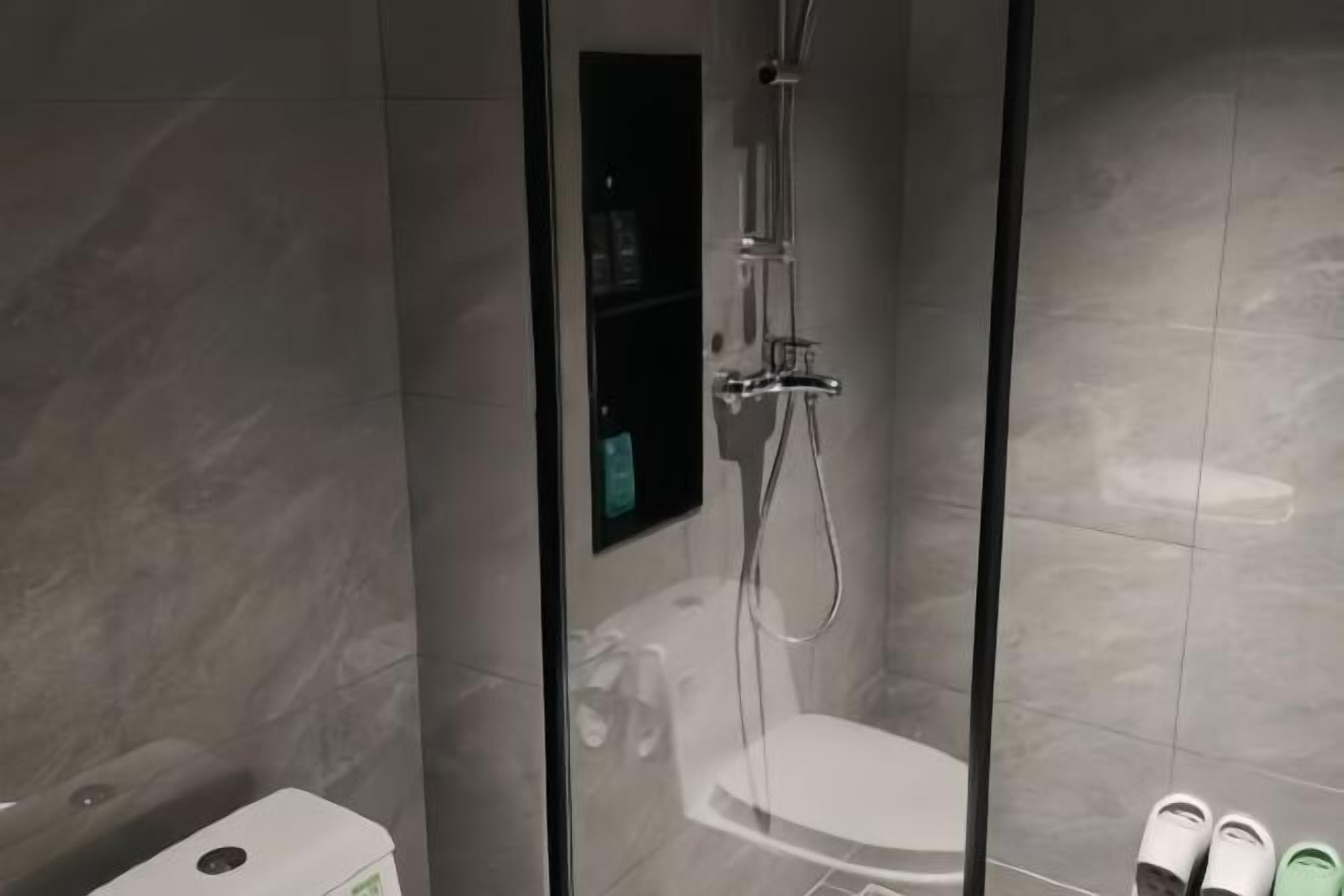 Un espace douche fermé par des parois de verre, doté d’un panneau de commande, de toilettes blanches et d’une paire de chaussons posés sur le sol.