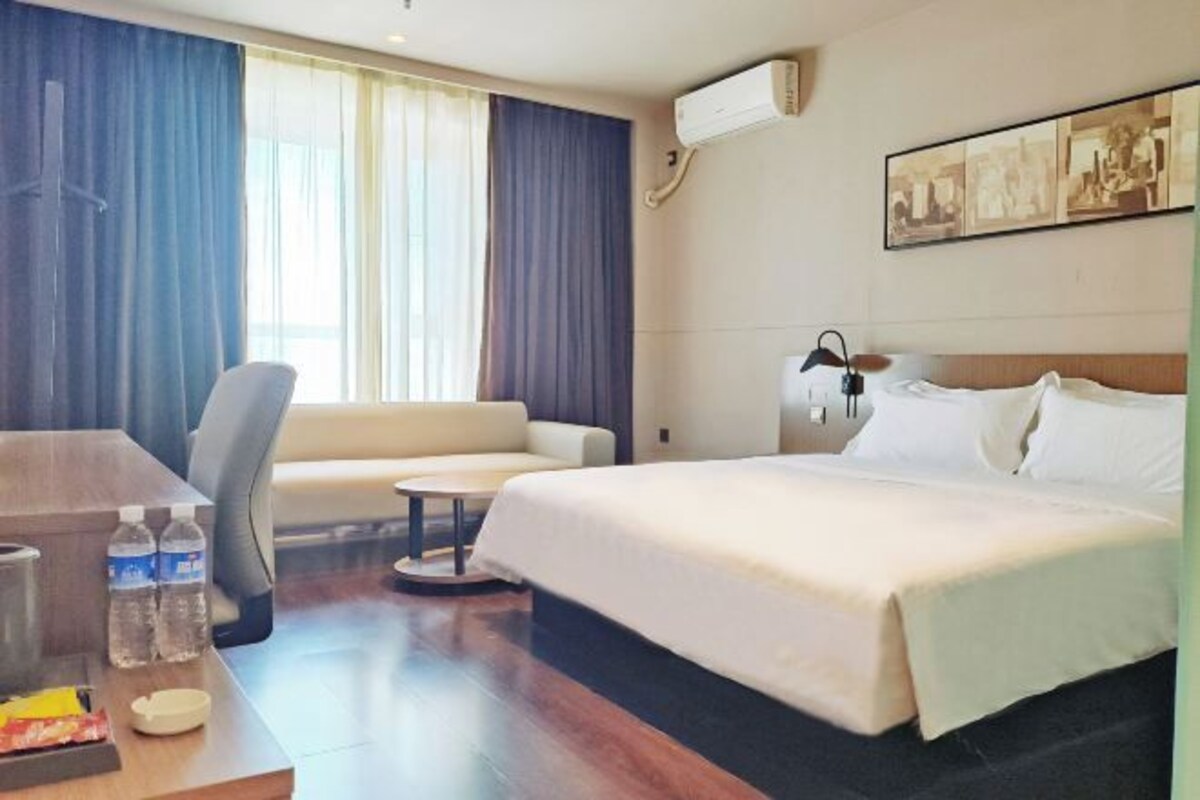 A hotel room with a bed, a sofa, a desk with water bottles, and a wall-mounted air conditioner.