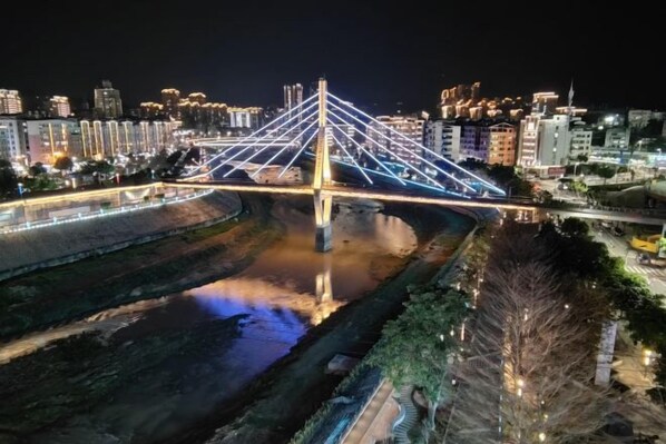 Vista nocturna de un puente con cables iluminados, un río debajo y edificios de la ciudad al fondo.