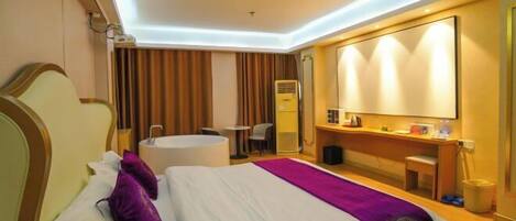A hotel room with a bed, a desk, a chair, a small table, a mirror, and a bathroom area.