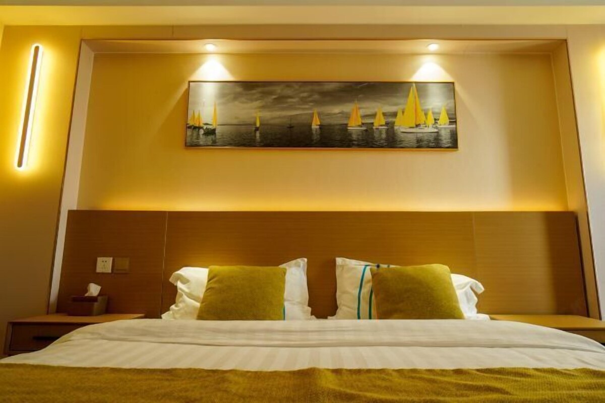 Habitación de hotel con cama, dos almohadas, una mesita de noche, un cuadro de veleros y una lámpara de pared.