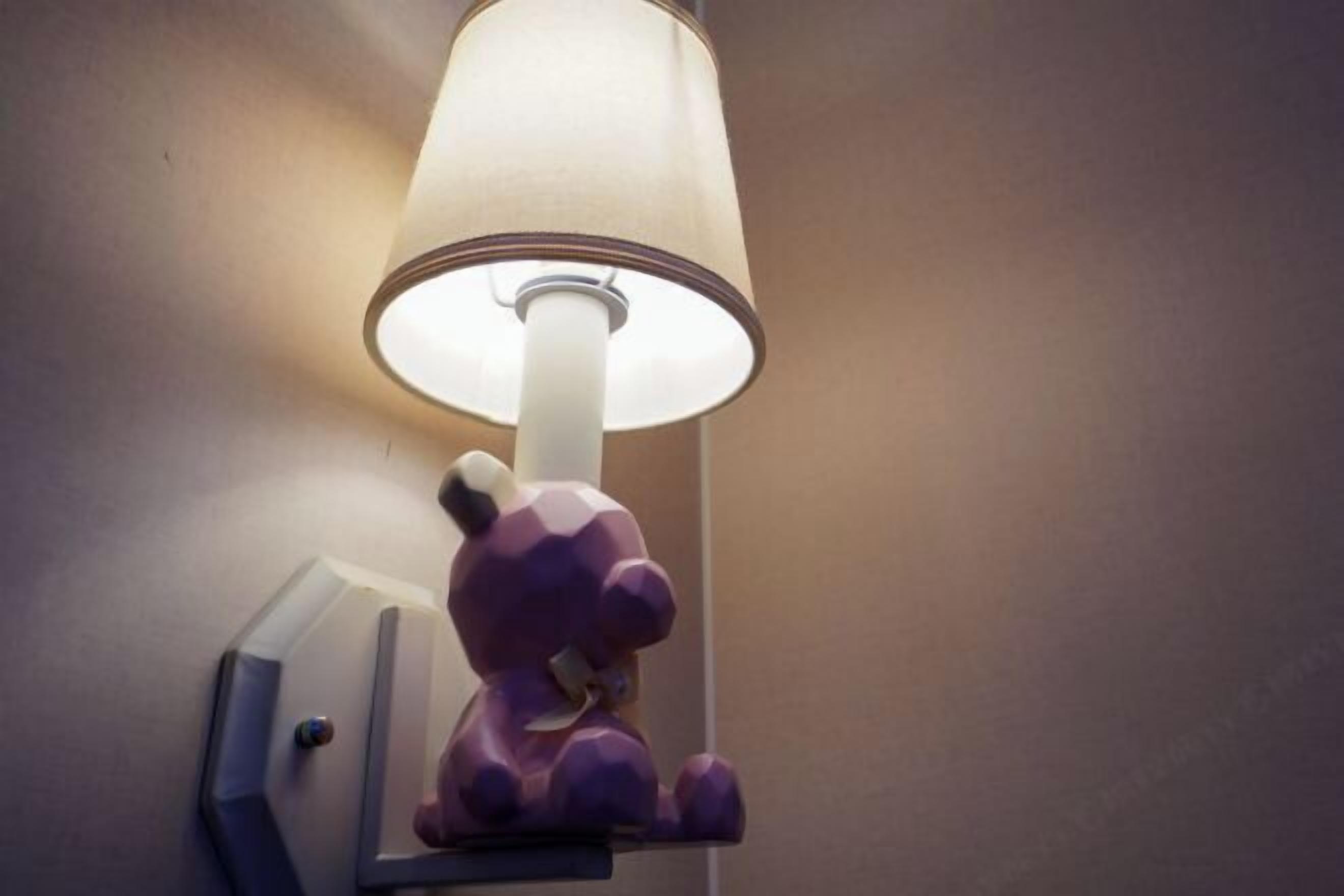 A wall-mounted lamp with a white shade and a purple bear-shaped nightstand.