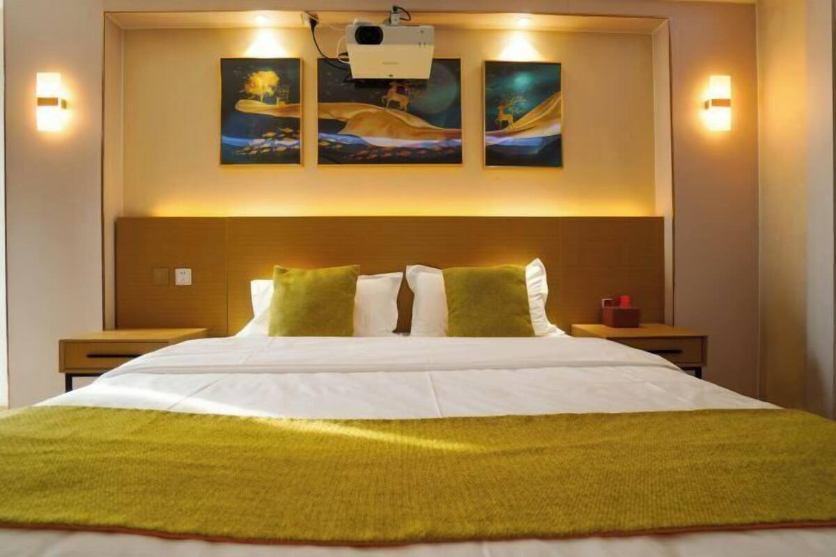 A hotel room with a large bed, a headboard with artwork, bedside lamps, and a projector mounted on the wall.