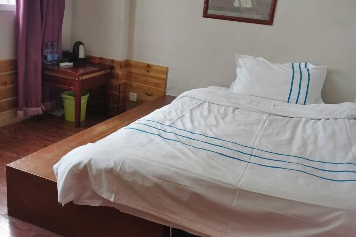 Una habitación con cama, un escritorio de madera, un cesto de basura verde y una ventana con cortinas moradas.