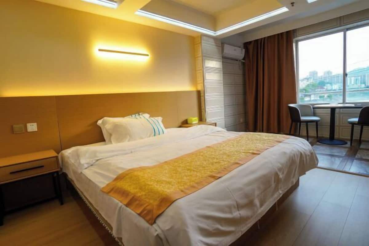 A hotel room with a large bed, a bedside table, a desk with two chairs, and a window with a city view.