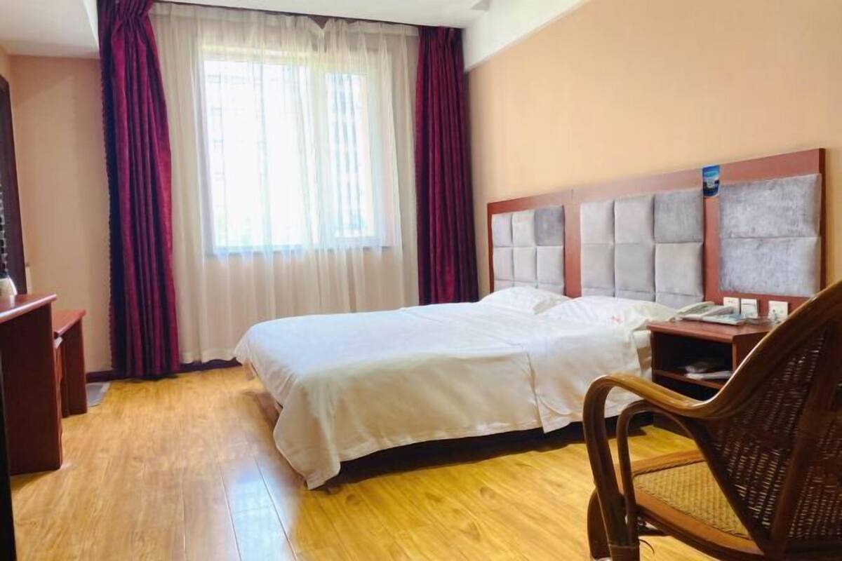Ein Hotelzimmer mit Bett, Holzboden, Vorhängen, einem Sessel und einem Nachttisch.