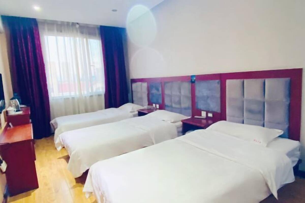 Ein Hotelzimmer mit drei Einzelbetten, einem Holztisch und einem Fenster mit Vorhängen.