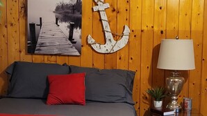 Ett sovrum med en väggpanel av trä, en säng med ett rött överkast, en tavla som föreställer en brygga och en vit ankardekoration.
