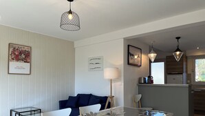 Una cocina moderna con una isla central, un comedor con mesa de vidrio y una sala de estar con sofá y lámpara.