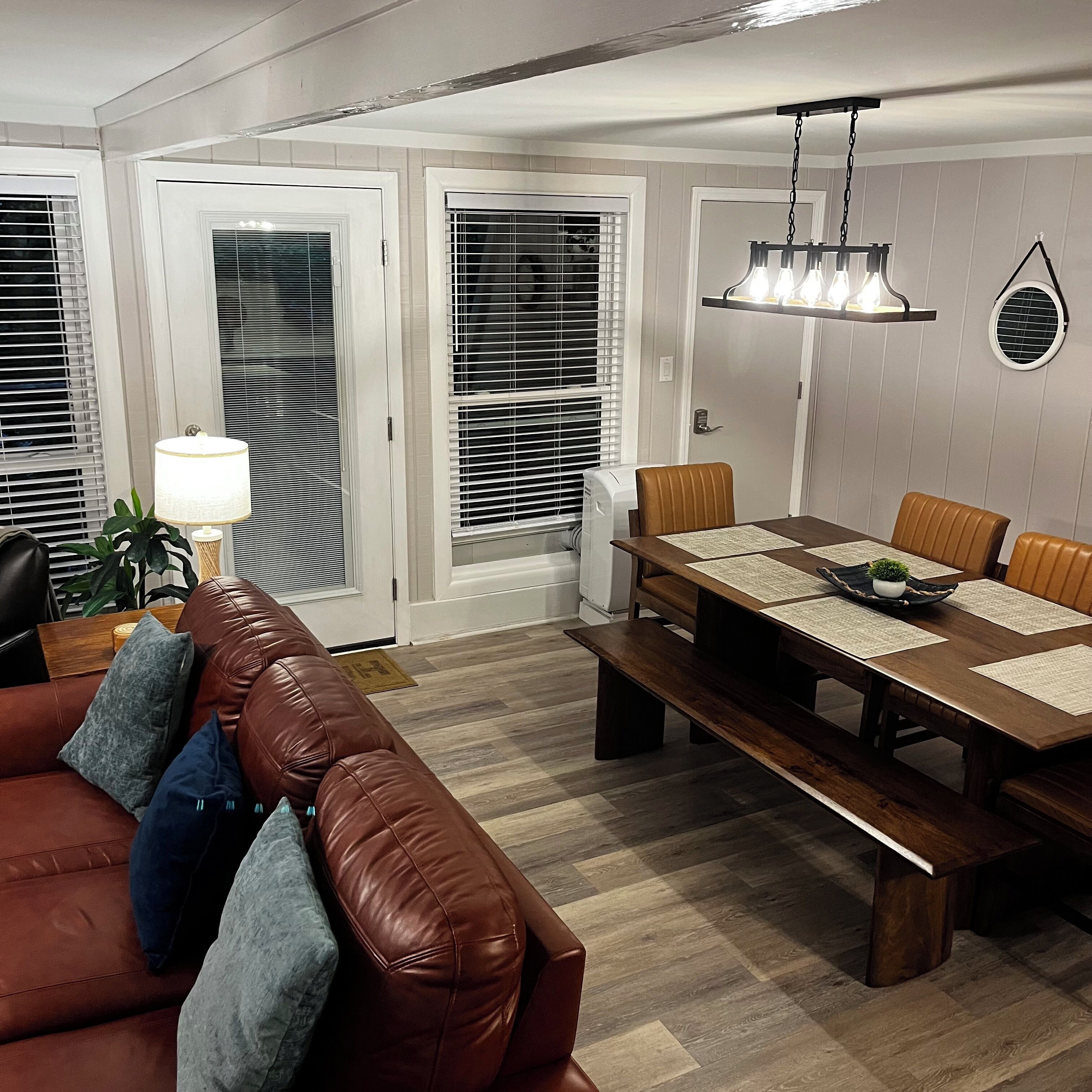 Ein Wohnzimmer mit braunem Ledersofa, einem Esstisch mit Stühlen und einer Deckenleuchte.
