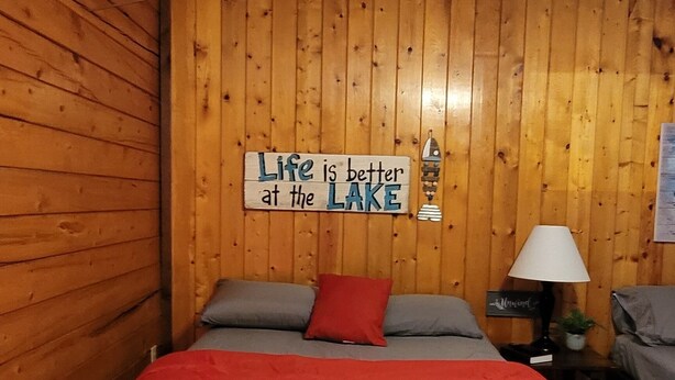 Une pièce aux murs lambrés, un lit recouvert d’une couverture rouge, des oreillers et une pancarte sur laquelle on peut lire : « La vie est plus belle au bord du lac ».