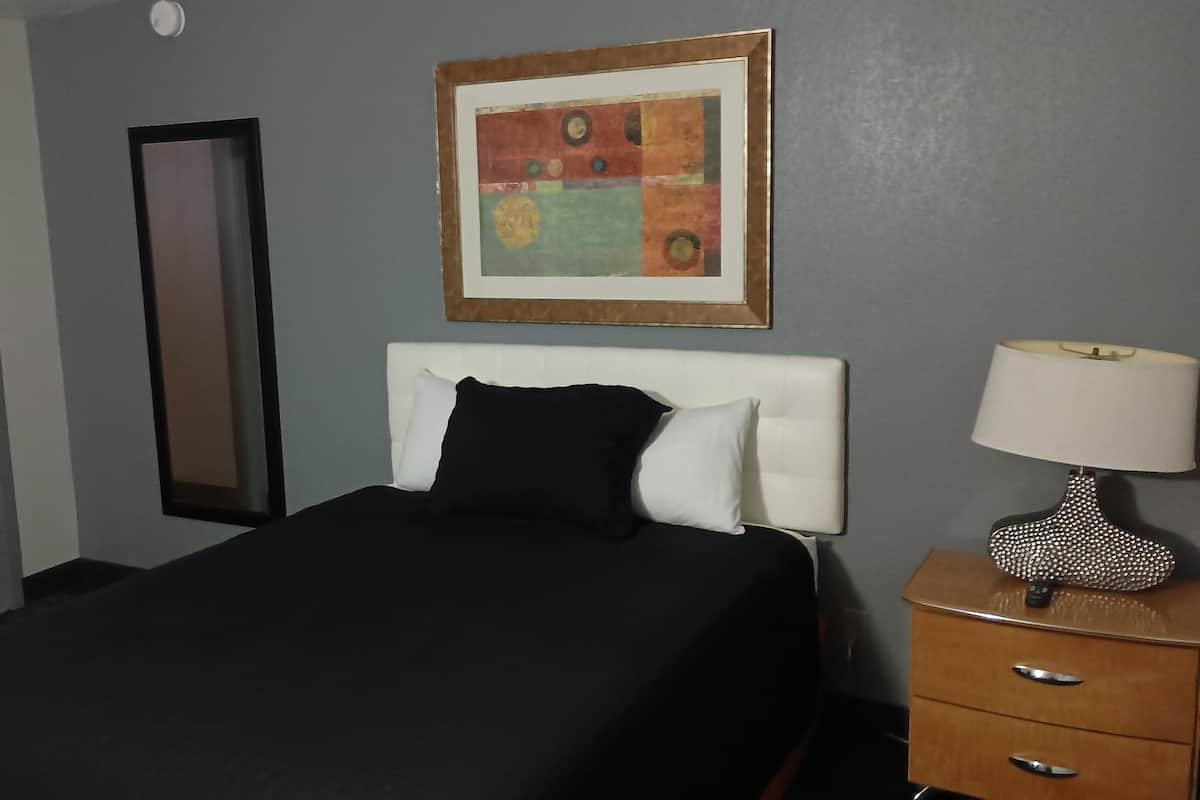 Ein Hotelzimmer mit einem Bett, einem Nachttisch mit Lampe, einem gerahmten Kunstwerk, einem Spiegel und einer Tür.