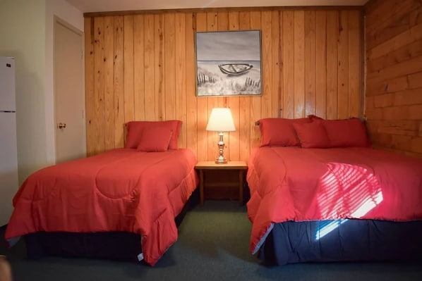 A room with two beds, each with red bedding, a wooden wall paneling, a nightstand with a lamp, and a painting of a boat on the wall. - #14 Shore Beats Work-Uncertain Lodge and Inn (Karnack)