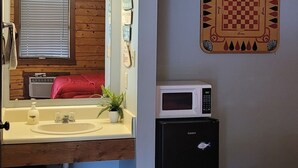 A compact room with a wooden vanity, a sink, a mirror, a small refrigerator, a red bed, and a wall-mounted chessboard. - #15 Off the Hook-Uncertain Lodge and Inn (Karnack)
